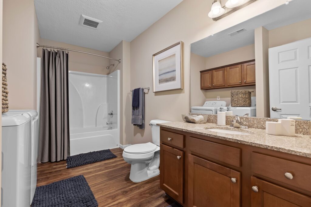 Bathroom with a bathtub and shower, toilet, sink with granite countertop, washer and dryer, wooden cabinets, and neutral decor.