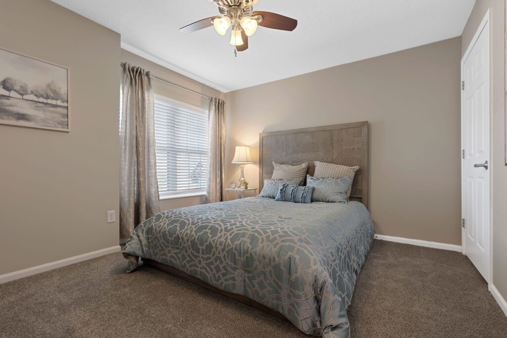 A neatly made bed with blue and beige bedding sits against a taupe wall in a carpeted bedroom, with a window, lamp, and ceiling fan visible.