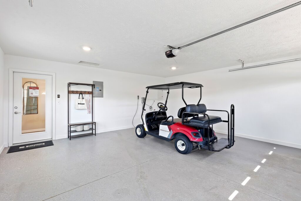 A red golf cart is parked in a clean, empty garage with white walls, a light gray floor, a door, and some shelves on the left side.
