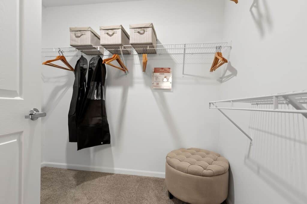 A walk-in closet with wire shelves, several wooden hangers, three fabric bins, a black garment bag, a tufted beige ottoman, and carpeted flooring.
