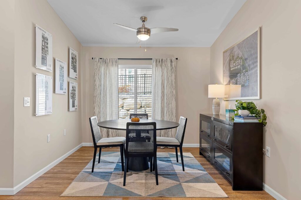 A small dining area with a round black table, four black chairs, a sideboard with a lamp and plant, wall art, and a window with sheer curtains.