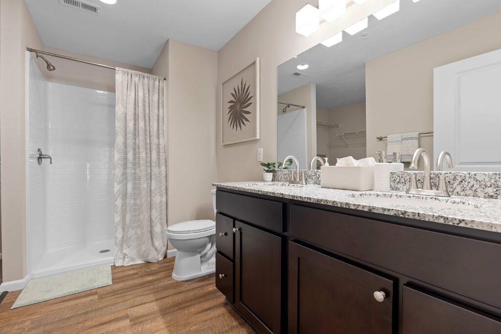 Modern bathroom with double sink vanity, granite countertop, dark cabinets, walk-in shower with curtain, toilet, large mirror, and wood-look flooring.