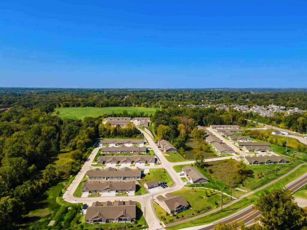Aerial view of a suburban neighborhood with rows of houses, roads, surrounding trees, and open grassy fields under a clear blue sky.