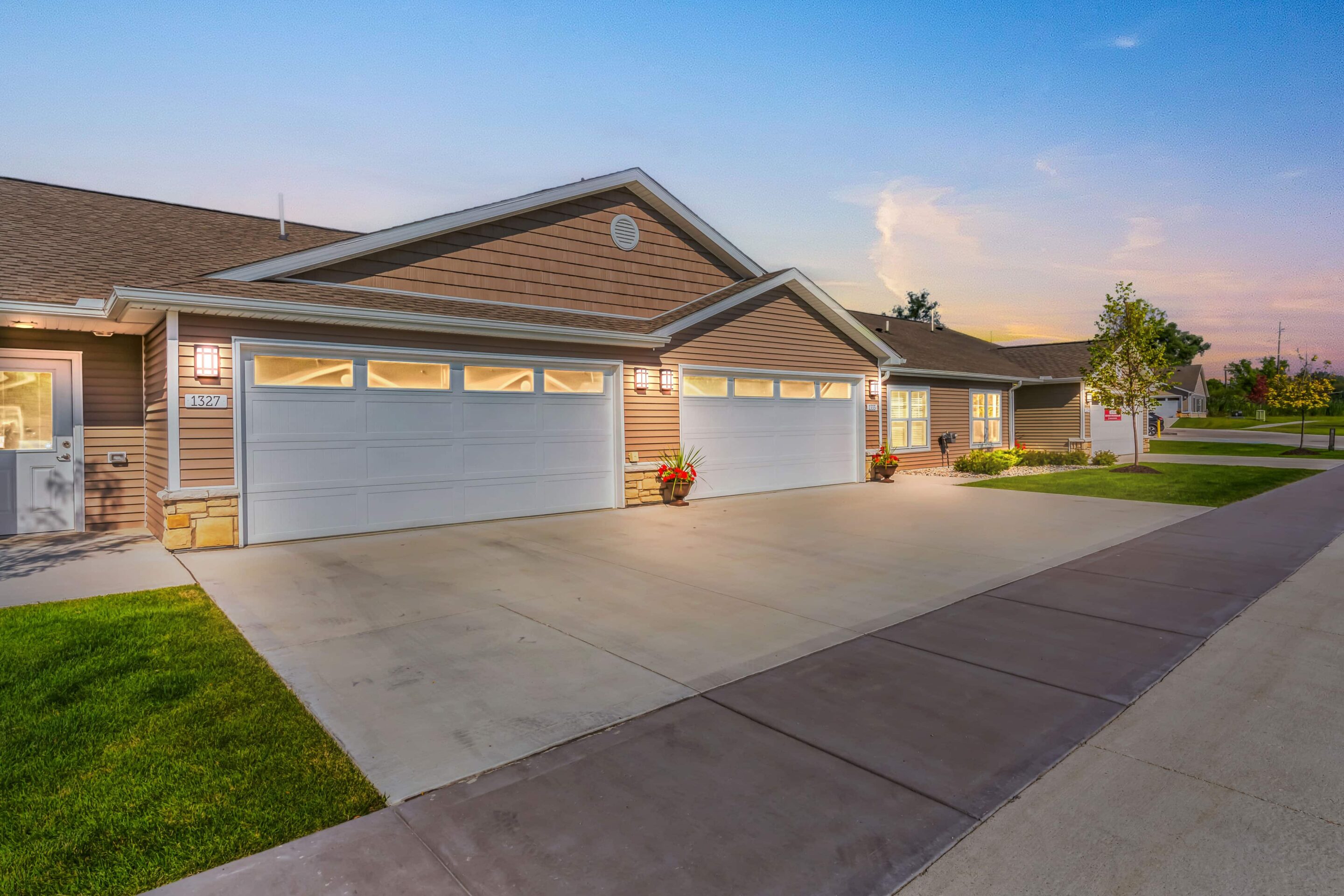 Single-story suburban houses with attached three-car garages, concrete driveways, green lawns, and outdoor lighting, set during sunset.