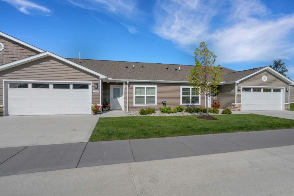 Single-story duplex with two garages, beige siding, white trim, small front yard, and a young tree, set against a blue sky with scattered clouds.