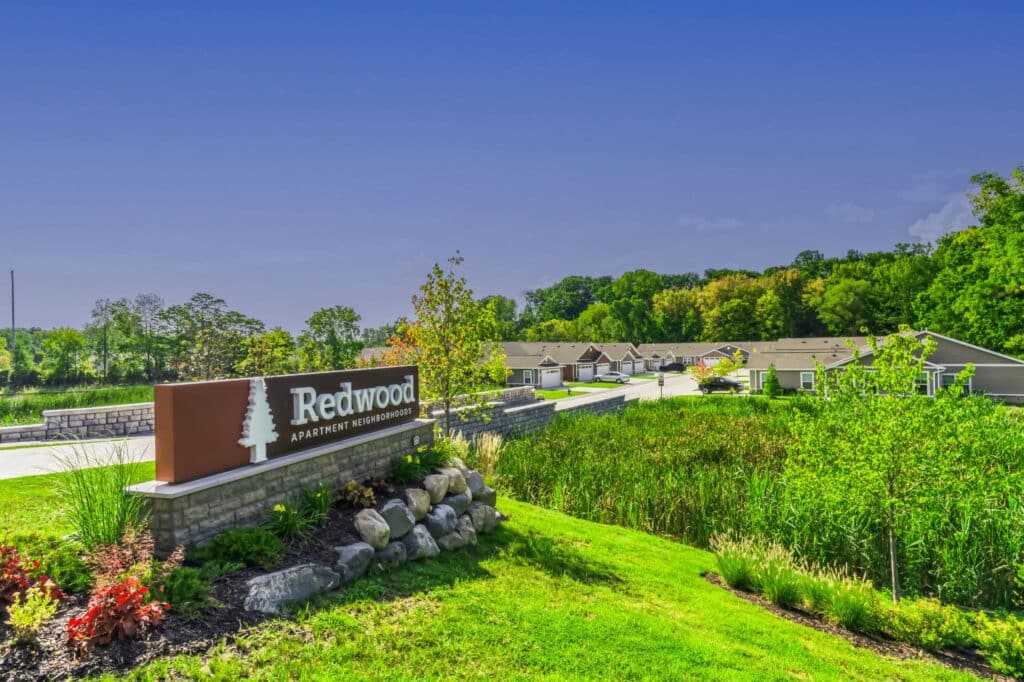 A sign reading “Redwood Apartment Neighborhoods” stands beside a landscaped area with rocks and grass, with residential buildings and trees in the background under a clear blue sky.