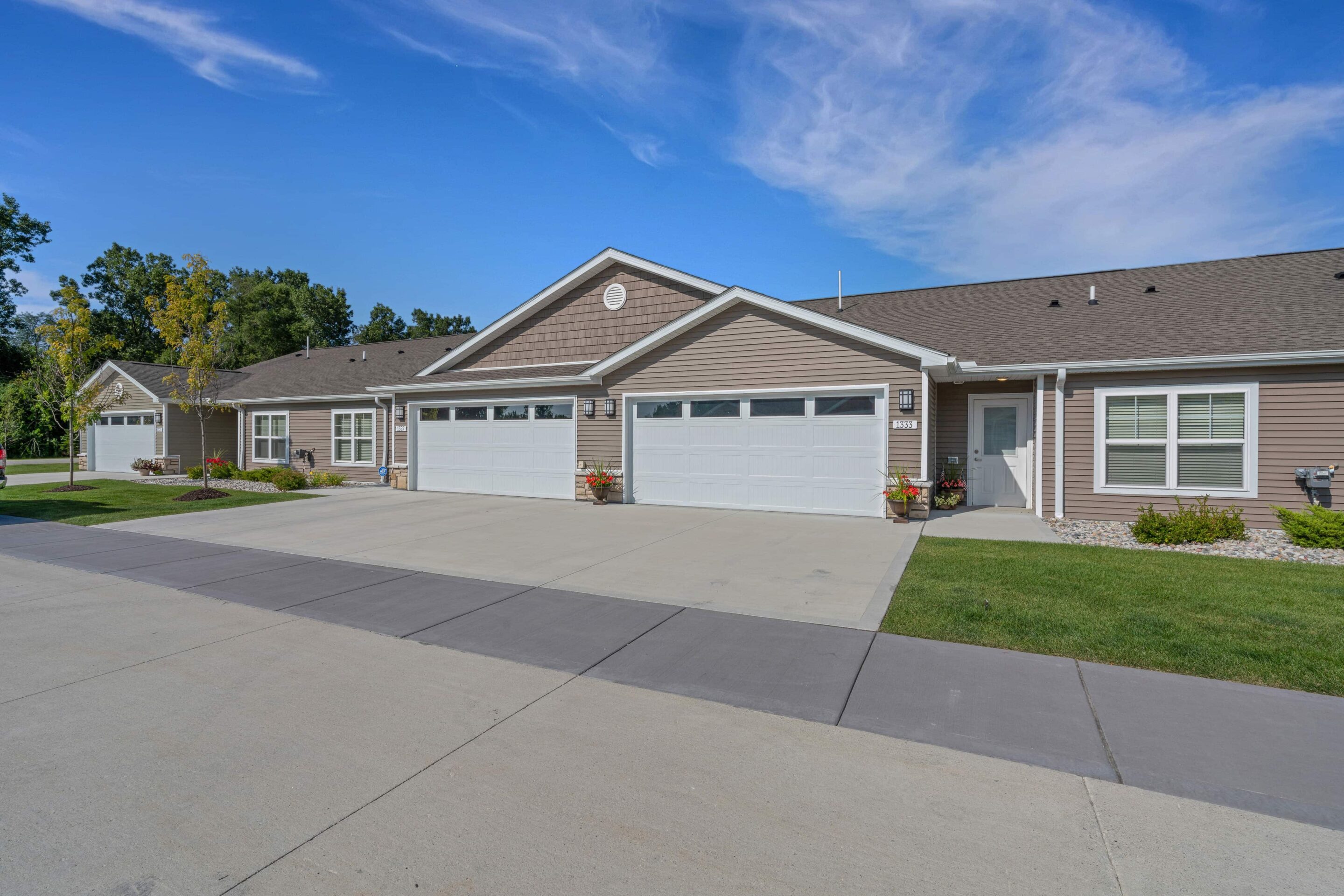 Single-story townhouse units with attached two-car garages, beige siding, and manicured lawns under a partly cloudy blue sky.