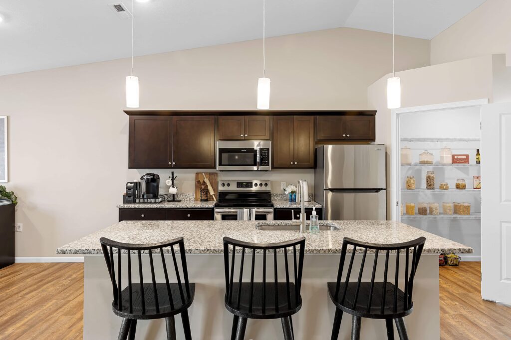 Modern kitchen with granite island, three black chairs, stainless steel appliances, dark wood cabinets, and an open pantry with organized jars on shelves.
