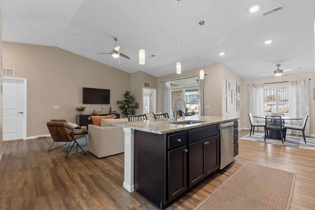 Open-concept living space with a kitchen island in the foreground, living area with sofa and TV, dining area by large windows, and neutral-colored walls and flooring.