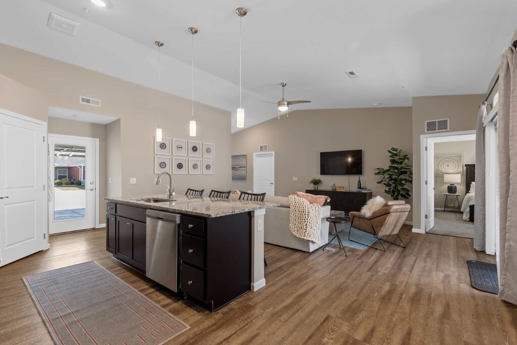 Open-concept living room and kitchen with island, pendant lights, wood flooring, neutral decor, and access to an outdoor area and bedroom.