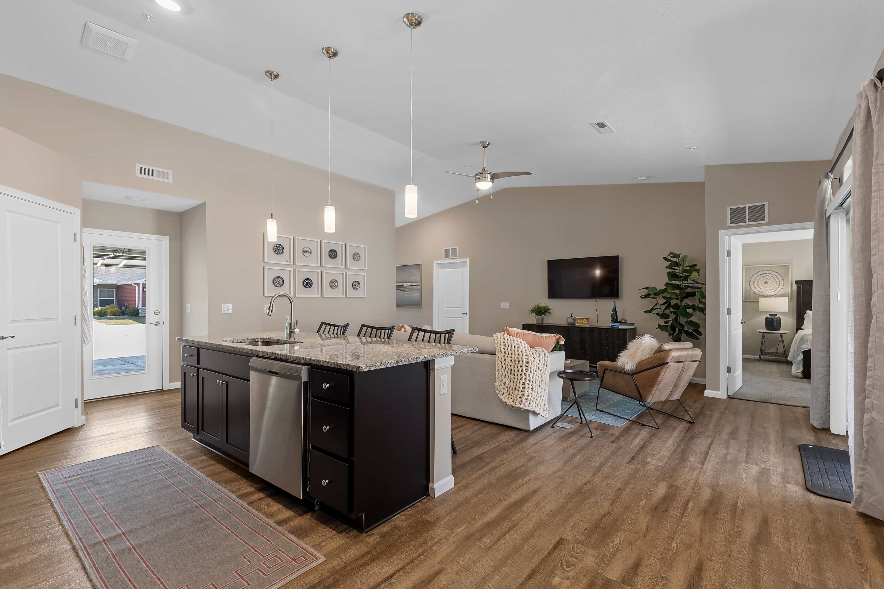 Open-concept living room and kitchen with island, pendant lights, wood flooring, neutral decor, and access to an outdoor area and bedroom.