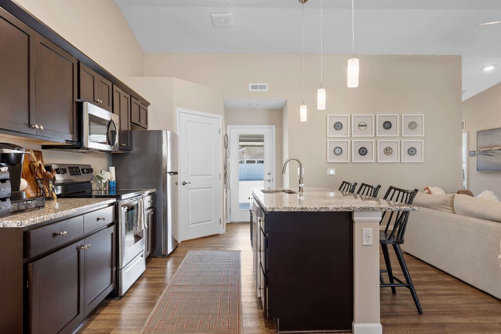 Modern kitchen with dark cabinets, stainless steel appliances, granite island with bar stools, and open view to a living area and entry door.