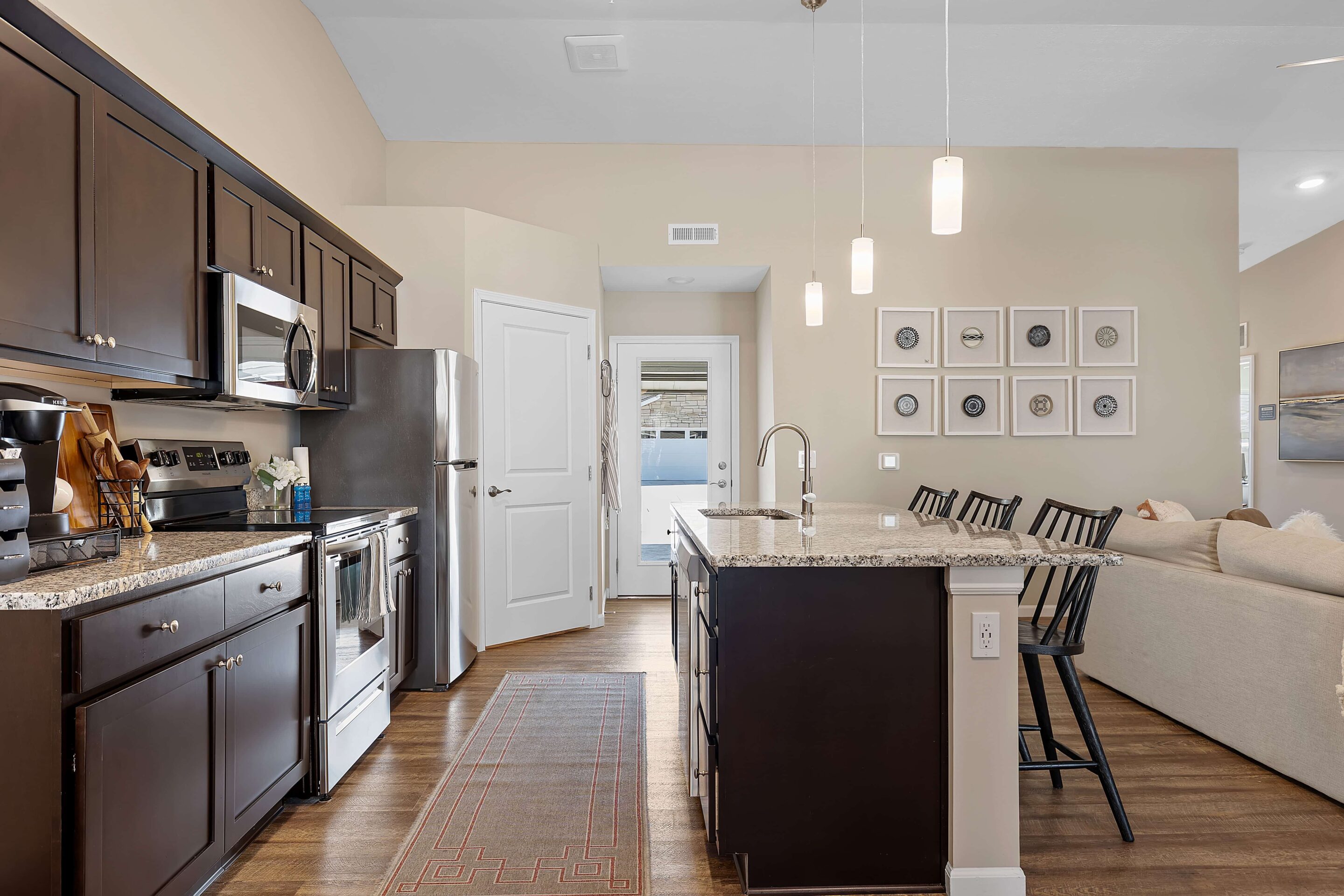 Modern kitchen with dark cabinets, stainless steel appliances, granite island with bar stools, and open view to a living area and entry door.