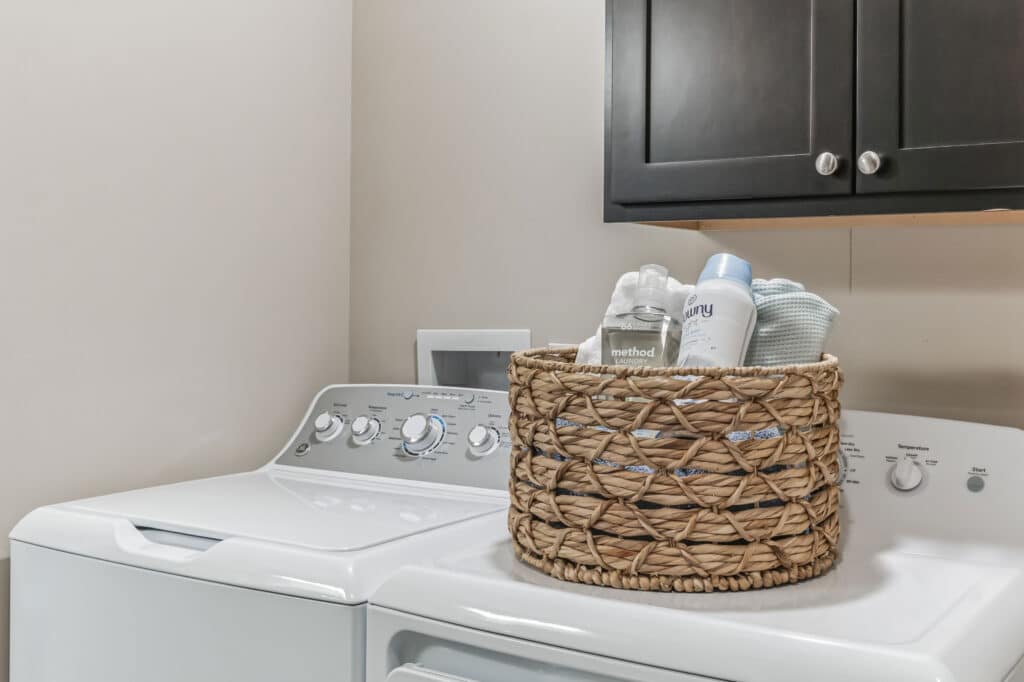A wicker basket with laundry supplies sits on top of a white washing machine next to a dryer in a laundry room with beige walls and dark cabinets.