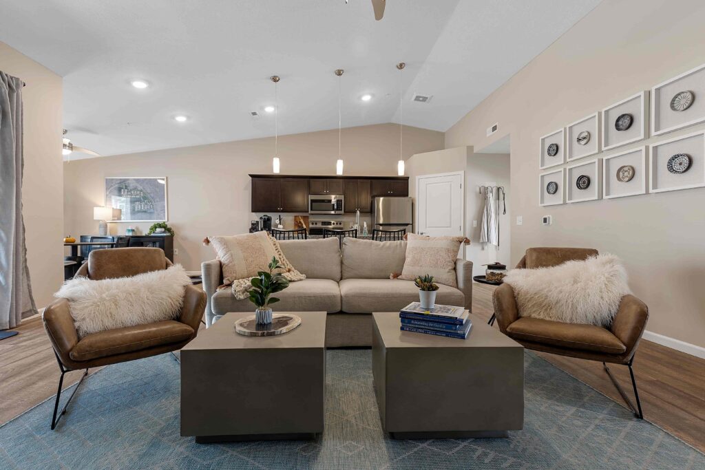 Modern living room with a beige sofa, two brown chairs, square coffee tables, and a kitchen in the background. Neutral tones and decorative wall art create a cozy, contemporary space.