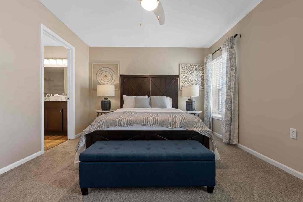 A neatly made bed with two lamps and decorative art above the headboard, a blue bench at the foot, and an open doorway to a bathroom in a neutral-toned bedroom.