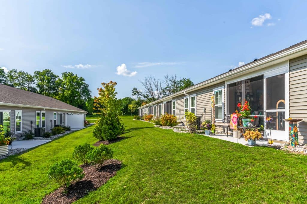 Row of single-story residential units with beige siding, landscaped lawns, and plants lining the walkway on a sunny day.