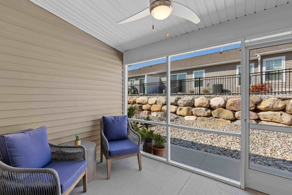 A screened-in patio with two blue cushioned chairs, a ceiling fan, potted plants, and a view of a rock retaining wall and neighboring building.