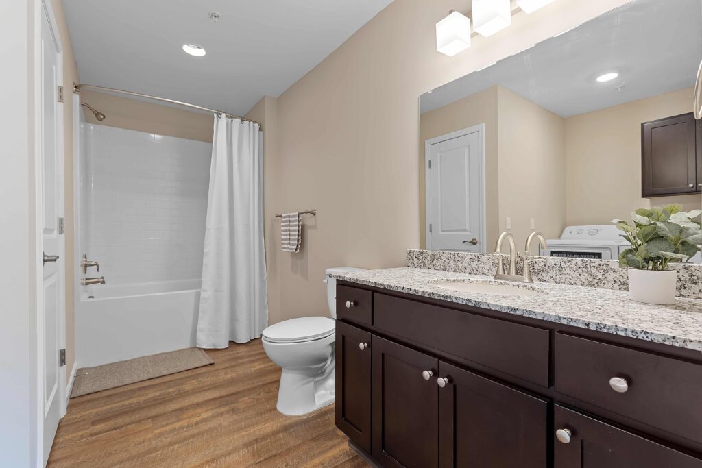 Modern bathroom with wood-look flooring, a white bathtub with shower curtain, granite countertop vanity, and a visible washing machine in the background.