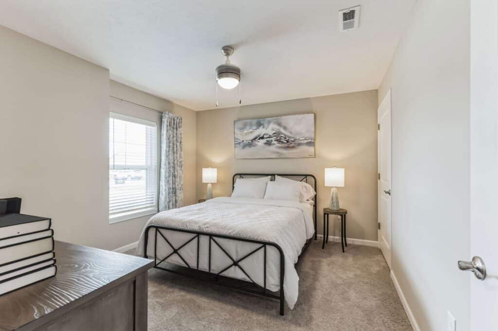 A bedroom with a bed, two side tables with lamps, a ceiling fan, a window with curtains, and a dresser with stacked books. Neutral wall colors and carpeted floor.