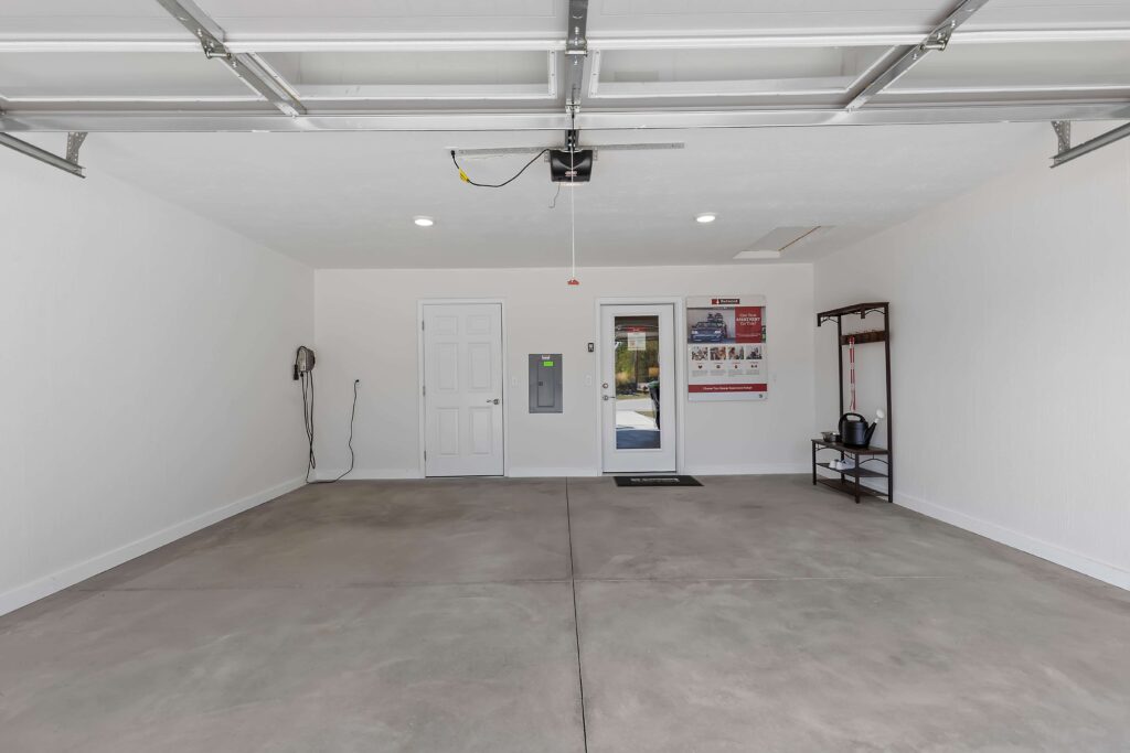 Clean, empty two-car garage with white walls, concrete floor, a door, electrical panel, shelf with items, and wall-mounted posters. Ceiling-mounted garage door opener visible.