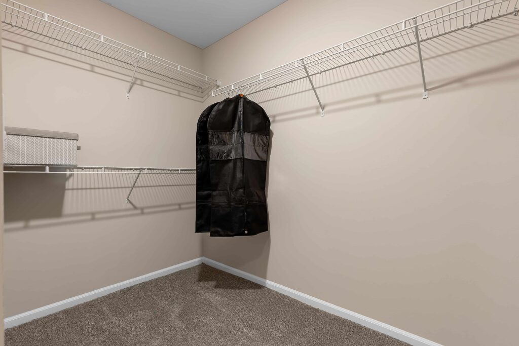 Empty walk-in closet with beige walls, wire shelving on two sides, one fabric garment bag hanging, and a storage box on the shelf. Carpeted floor.