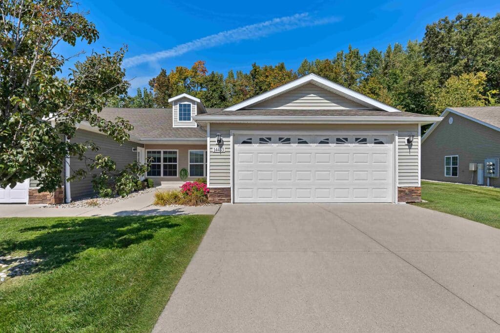 Single-story suburban house with attached two-car garage, light siding, manicured lawn, flowering shrubs, and trees in the background under a clear blue sky.