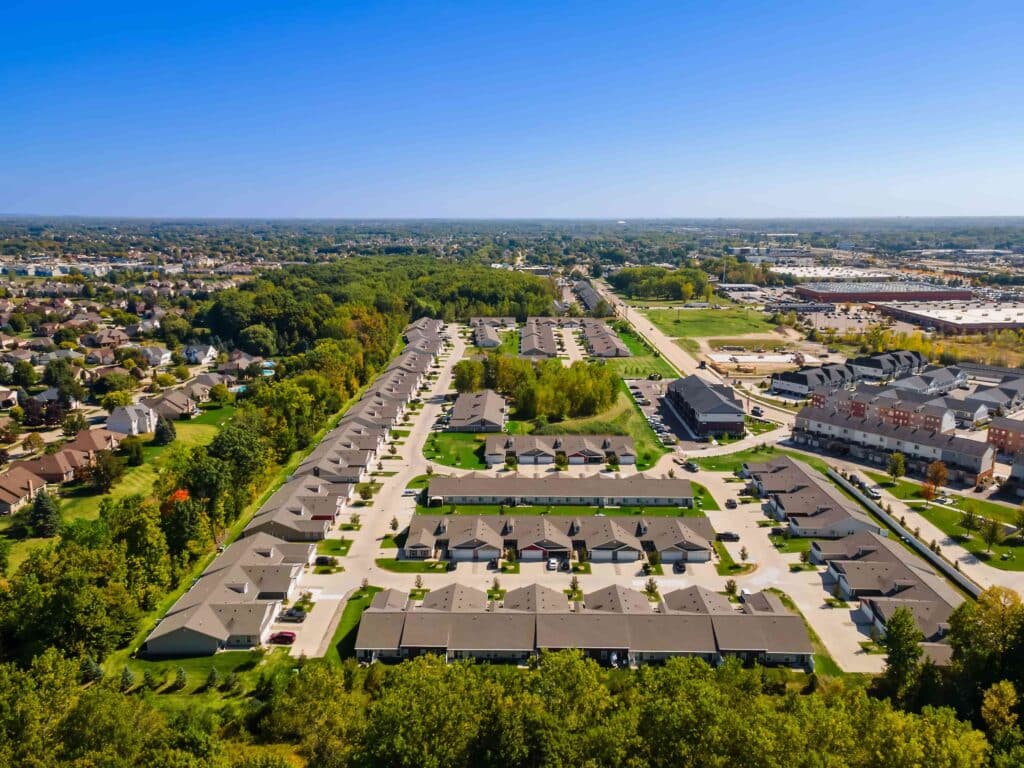 Aerial view of a suburban residential neighborhood with rows of houses, green lawns, trees, and roads under a clear blue sky.