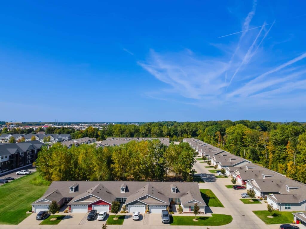 Aerial view of a suburban neighborhood with rows of single-family houses, tree-lined streets, and a forested area in the background under a clear blue sky.