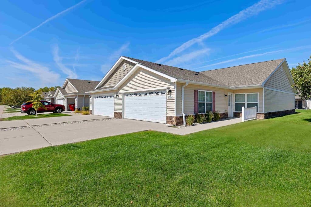 Single-story, beige-sided duplex with three-car garages, concrete driveways, and well-maintained green lawns under a blue sky with light clouds.