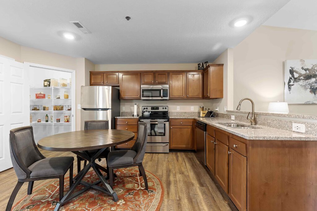 Modern kitchen with stainless steel appliances, wooden cabinets, round dining table with four chairs, open pantry, and granite countertops.