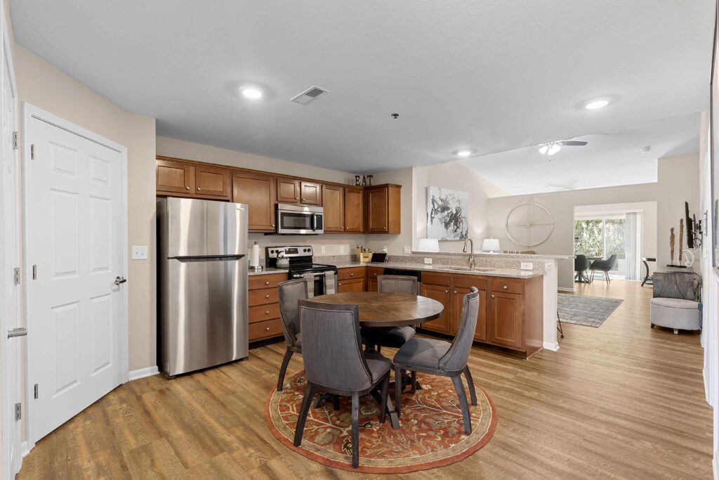Open-plan kitchen and dining area with stainless steel appliances, wooden cabinets, round dining table with four chairs on a patterned rug, and a view into the living room.