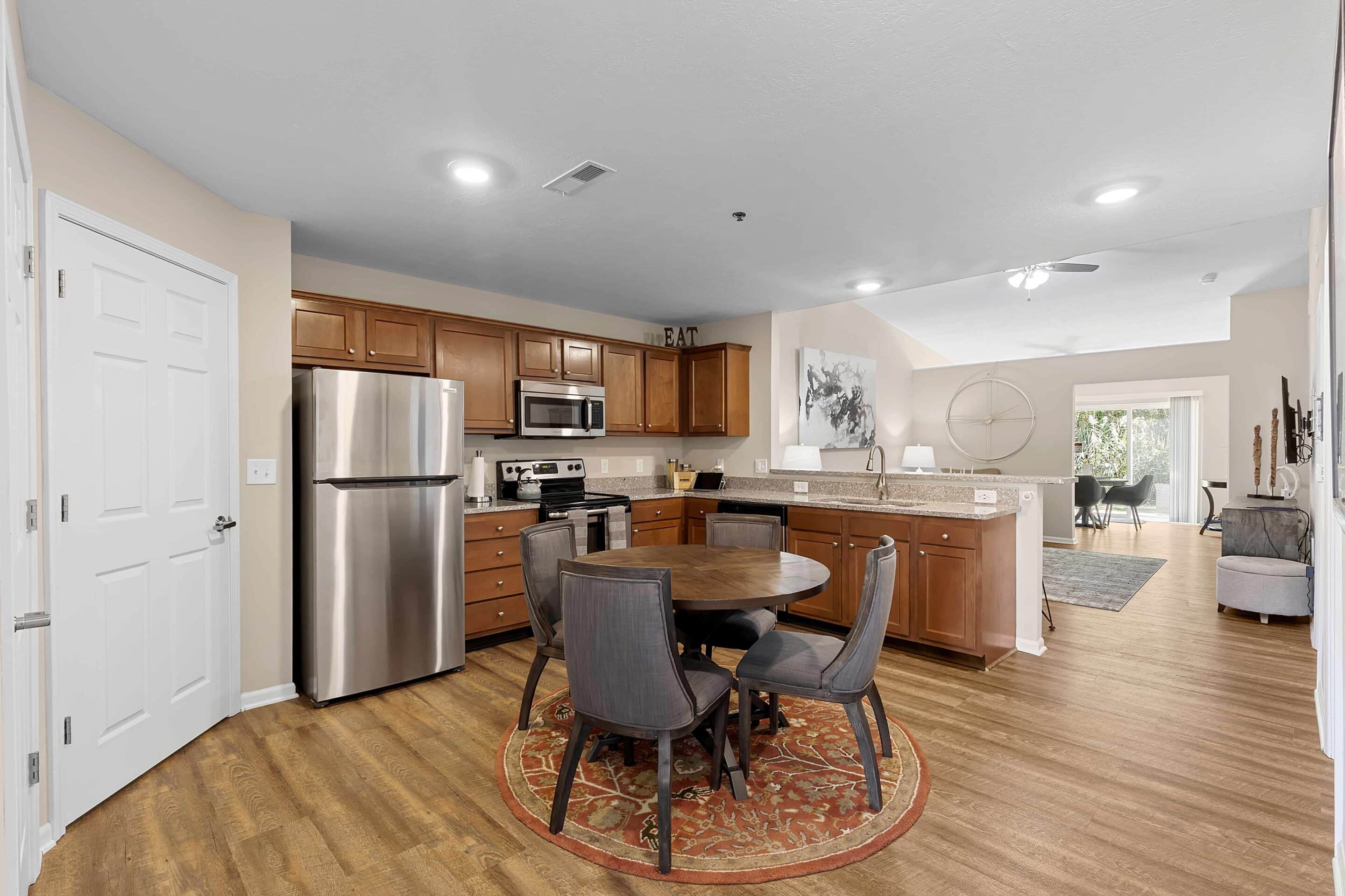 Open-plan kitchen and dining area with stainless steel appliances, wooden cabinets, round dining table with four chairs on a patterned rug, and a view into the living room.