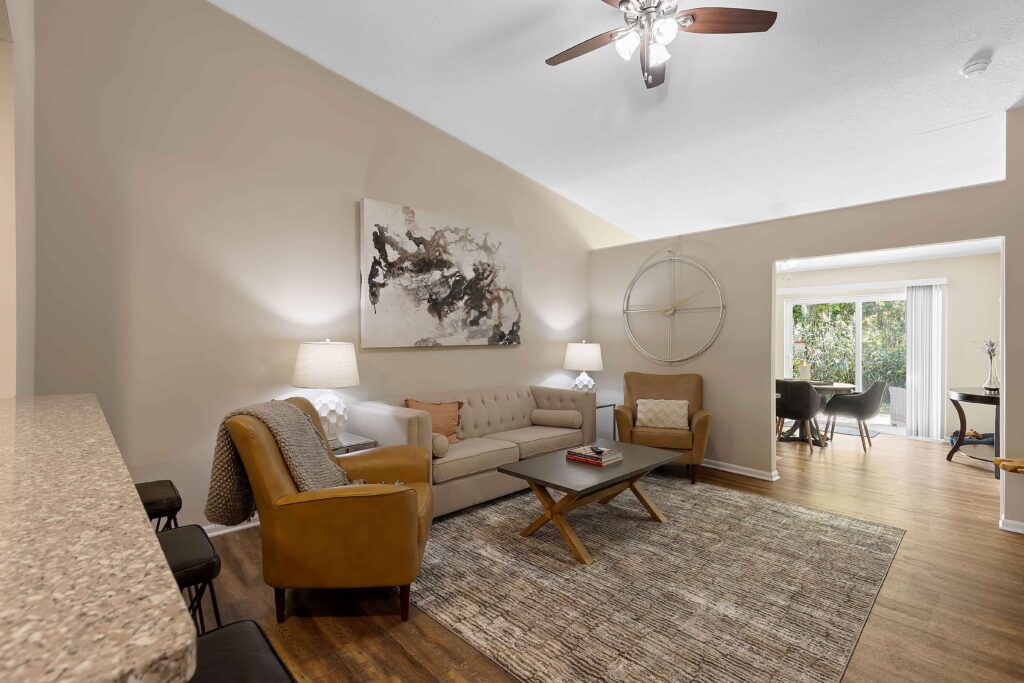 A modern living room with a beige sofa, two armchairs, a coffee table, a large wall clock, abstract artwork, and view into a dining area with natural light.