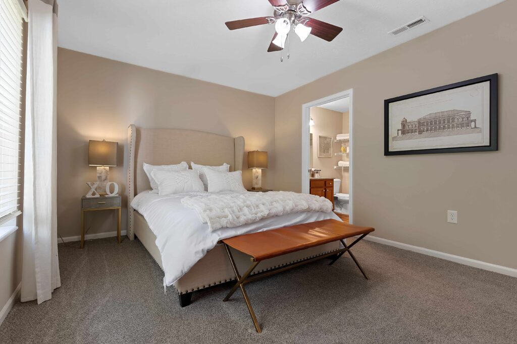 A neatly made bed with white bedding, flanked by two bedside tables and lamps, sits in a carpeted bedroom with beige walls, a ceiling fan, and an adjacent bathroom.