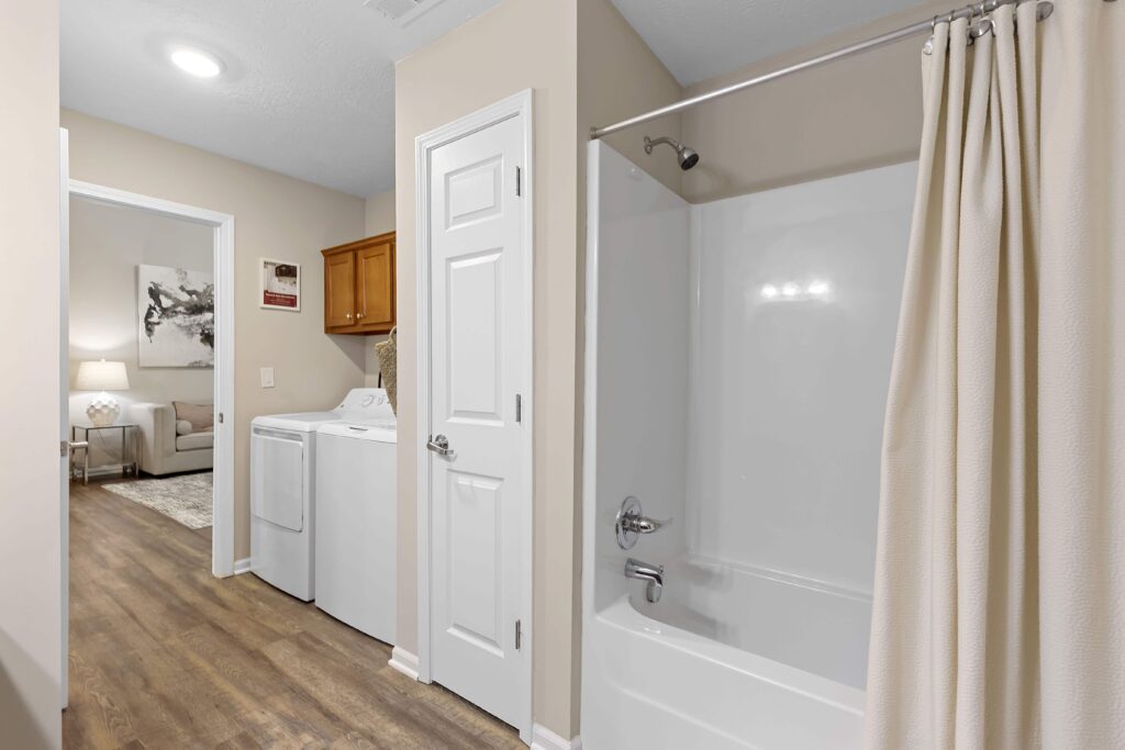 A bathroom with a white bathtub and shower curtain, adjacent to a laundry area with a washing machine and dryer, and a door leading to a living room.