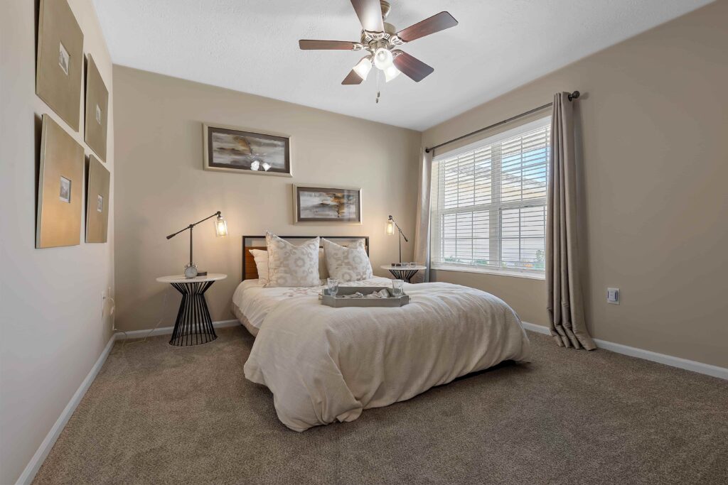 A neatly made bed with neutral bedding is centered in a carpeted bedroom, flanked by side tables and lamps, with wall art, a ceiling fan, and sunlight streaming through a window.