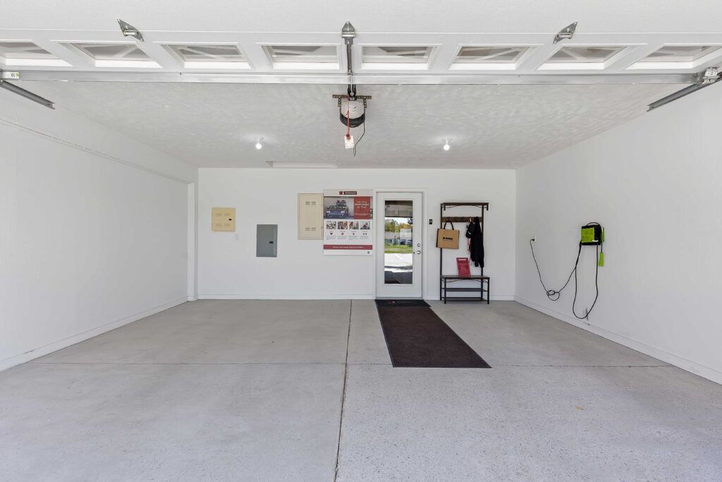 Clean, empty two-car garage with white walls, a door at the back, wall-mounted electrical panels, a coat rack, floor mat, and an electric vehicle charger on the right wall.