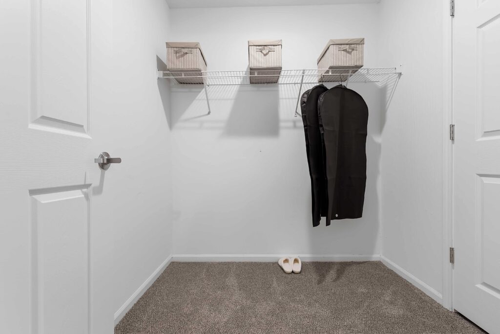 A walk-in closet with a wire shelf holding three storage boxes, two garment bags hanging, and a pair of white slippers on the carpeted floor.