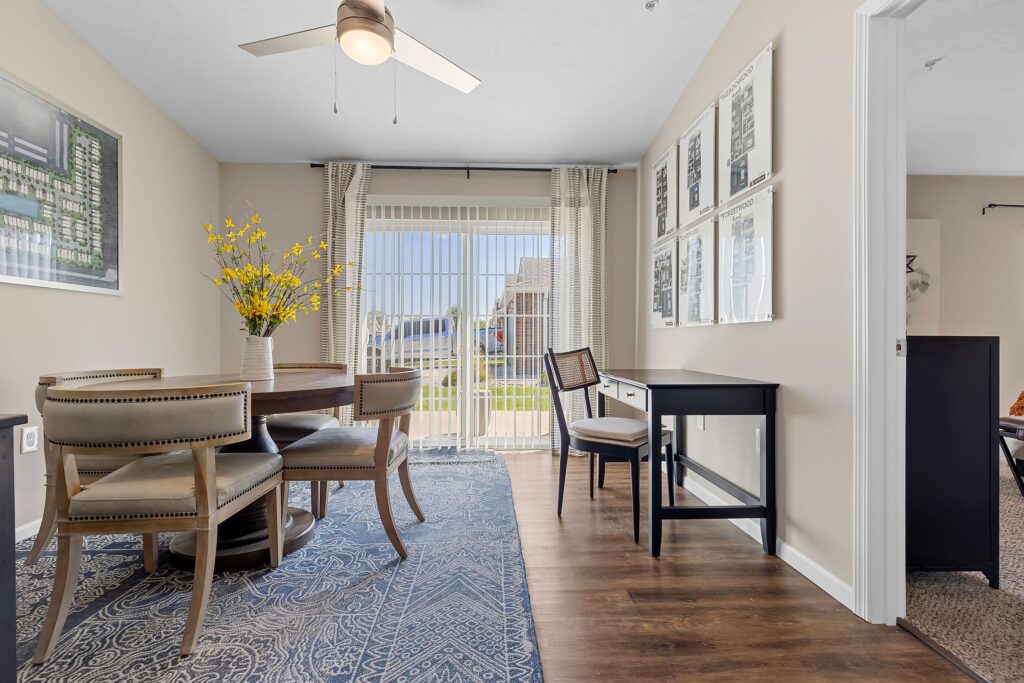 A dining area with a round table, four chairs, a vase of yellow flowers, a desk, and large sliding glass doors letting in natural light.