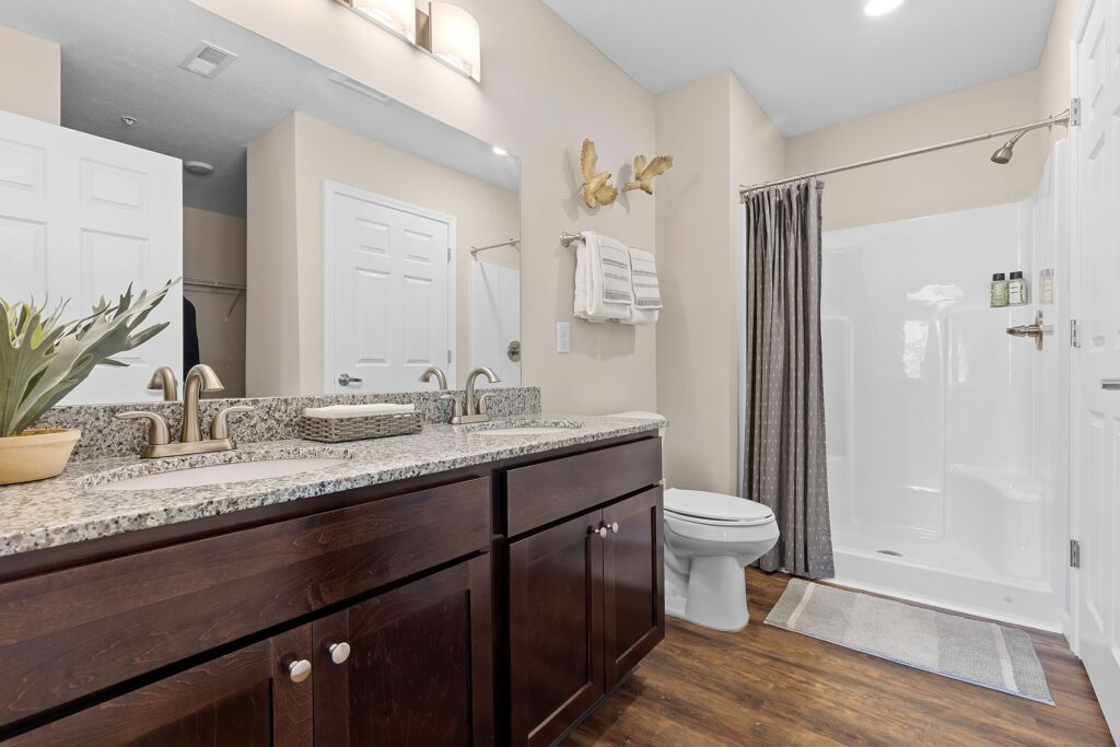 Modern bathroom with double sink vanity, granite countertop, dark wood cabinets, toilet, walk-in shower with gray curtain, and light-colored walls and flooring.