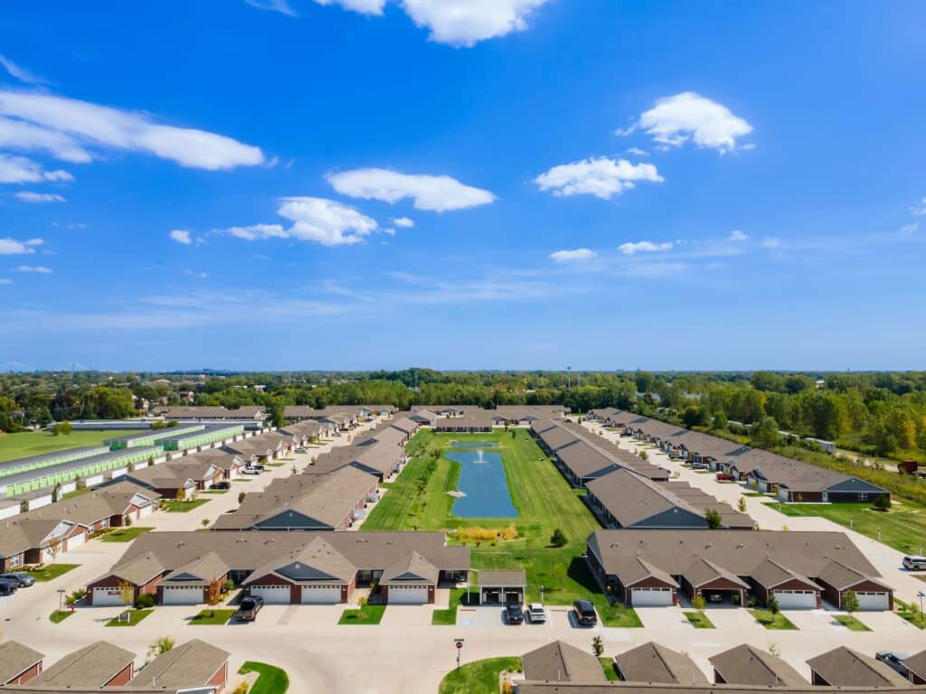 Aerial view of a residential neighborhood with rows of single-story houses arranged around a central grassy area with a pond, under a blue sky with scattered clouds.