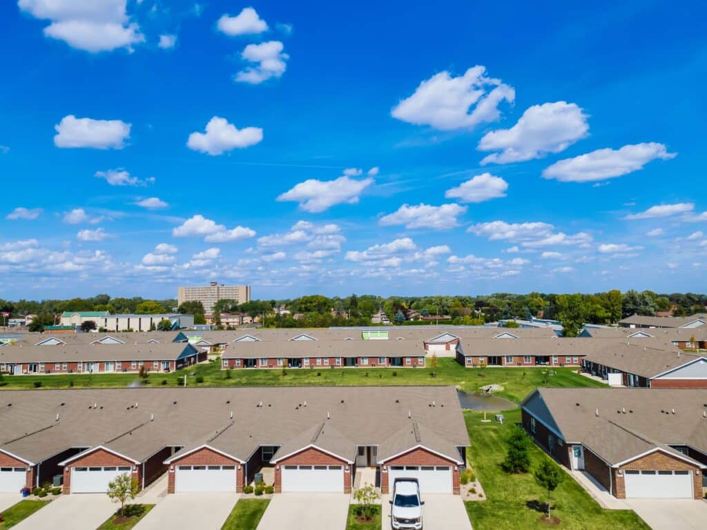 Aerial view of a suburban neighborhood with rows of single-story homes, driveways, green lawns, and a clear blue sky with scattered clouds.