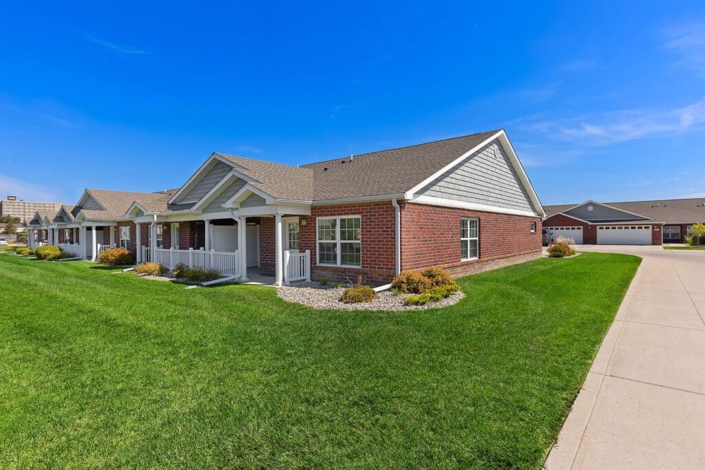Single-story brick house with white trim, a covered porch, and well-maintained lawn under a clear blue sky. Garage buildings are visible in the background.