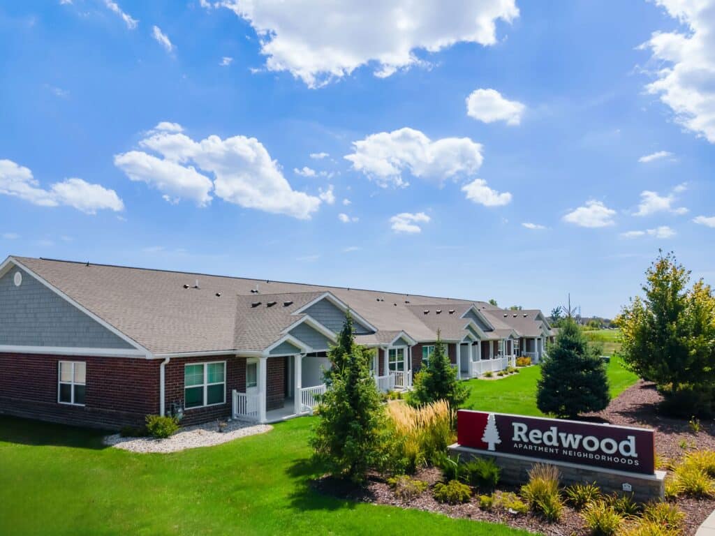 Row of single-story apartment homes with a "Redwood Apartment Neighborhoods" sign in front, surrounded by green grass, trees, and landscaped bushes under a blue sky with clouds.