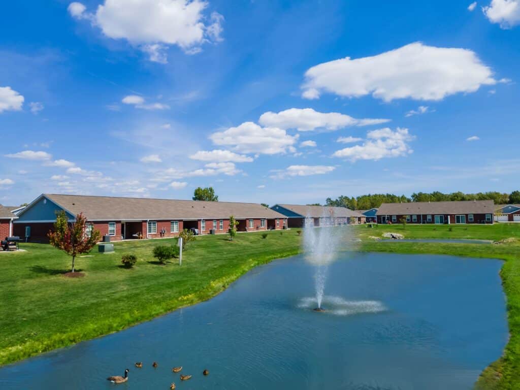 A small pond with a fountain, surrounded by grass, ducks swimming, and single-story residential buildings in the background under a blue sky with scattered clouds.