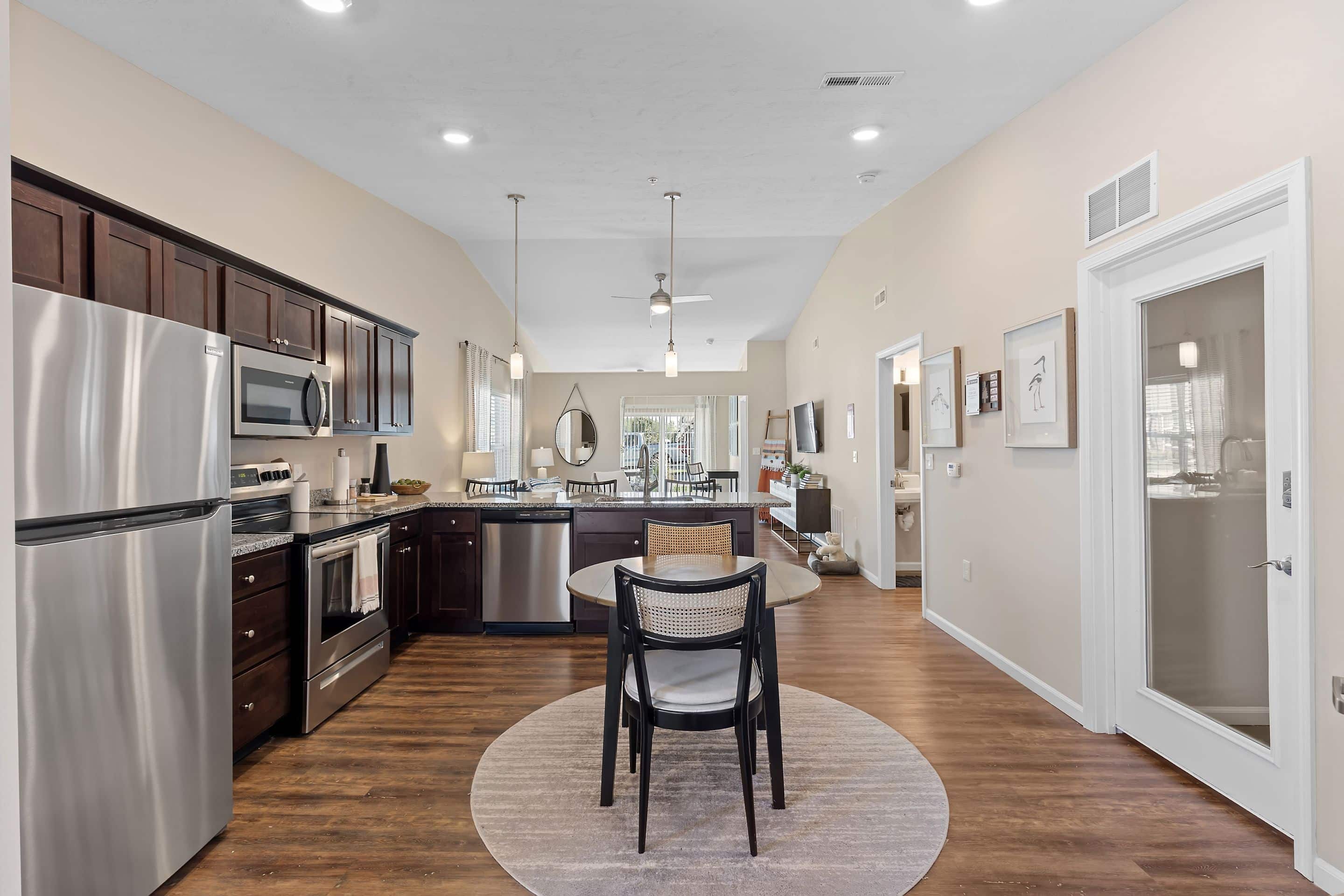 Modern kitchen and dining area with stainless steel appliances, dark wood cabinets, round dining table, and open view into a living room with a TV and windows.