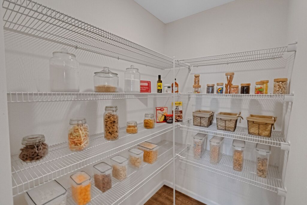 Wire shelves in a pantry hold jars and containers filled with various dry food items, including snacks, cereals, pasta, and seasonings, all neatly organized.