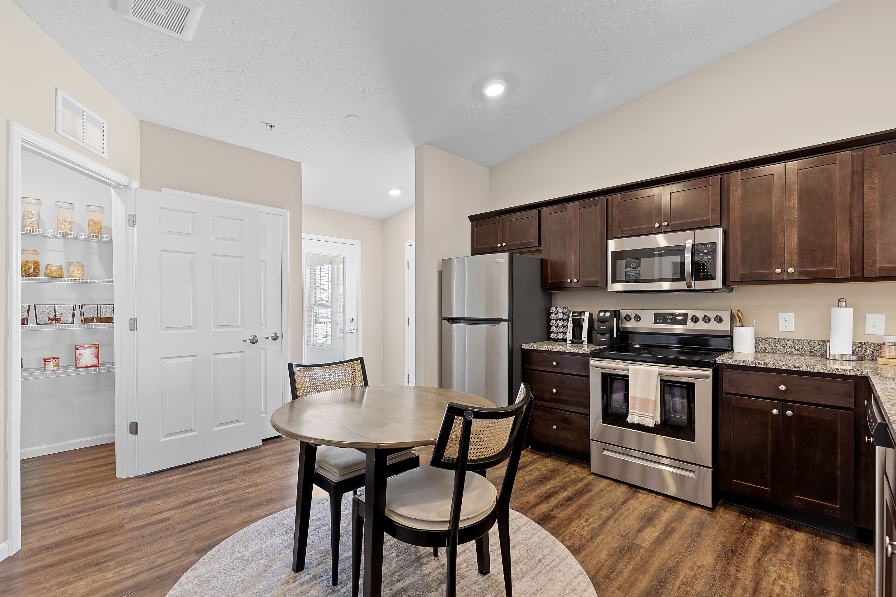 Modern kitchen with dark wood cabinets, stainless steel appliances, a round dining table with two chairs, and an open pantry stocked with jars and containers.