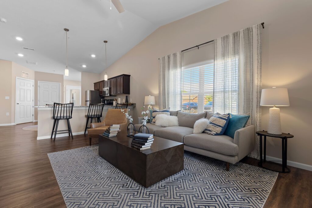 Modern living room with a gray sofa, patterned rug, coffee table, kitchen area with barstools, and large window with white curtains letting in natural light.
